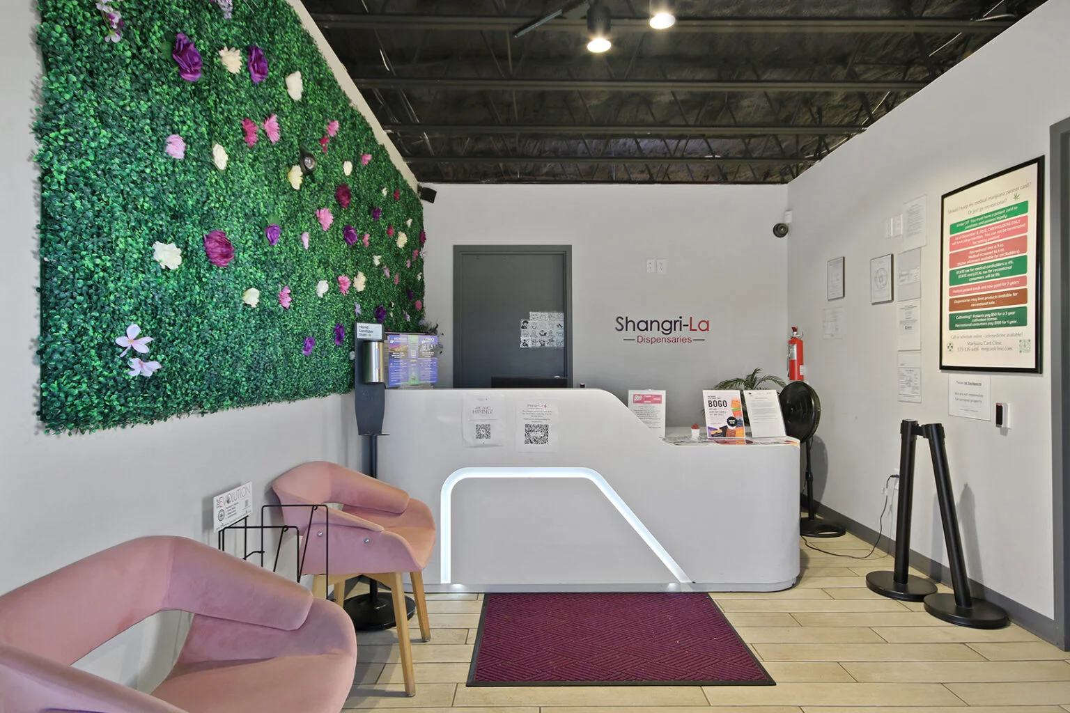 Modern dispensary reception area featuring a vibrant green wall with artificial flowers, sleek white reception desk with illuminated accents, and cozy pink seating. Informational signage and promotional materials are displayed prominently.