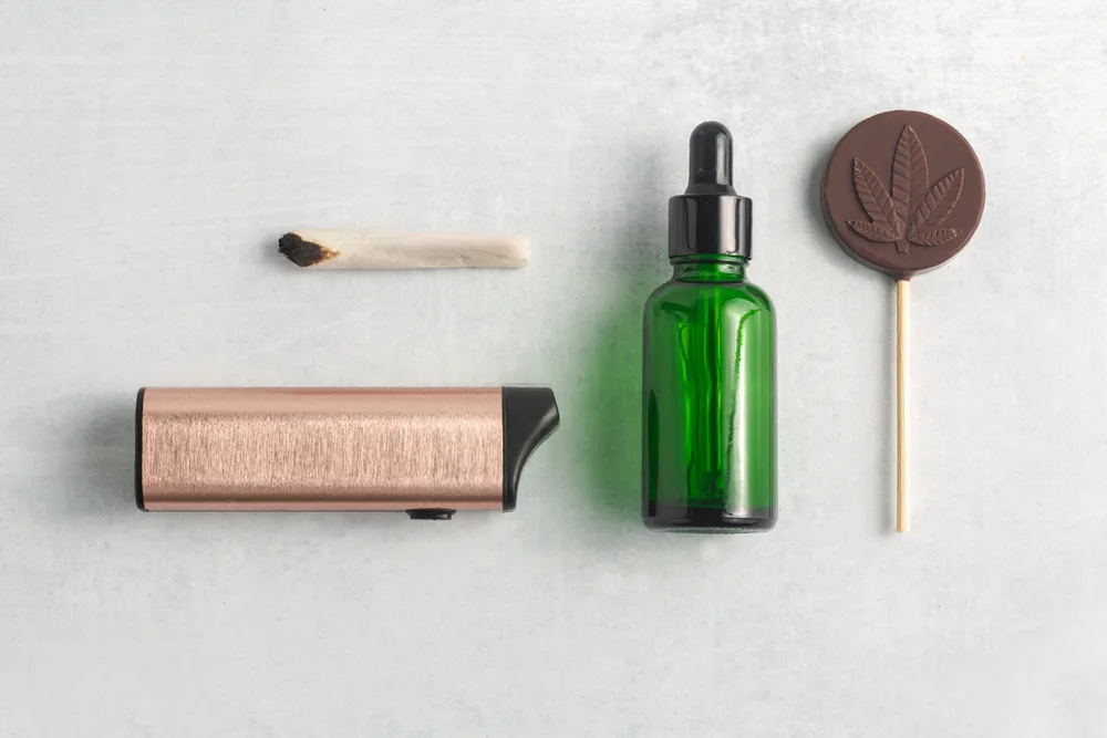 Various cannabis products including a joint, vape pen, dropper bottle, and chocolate lollipop with a cannabis leaf imprint